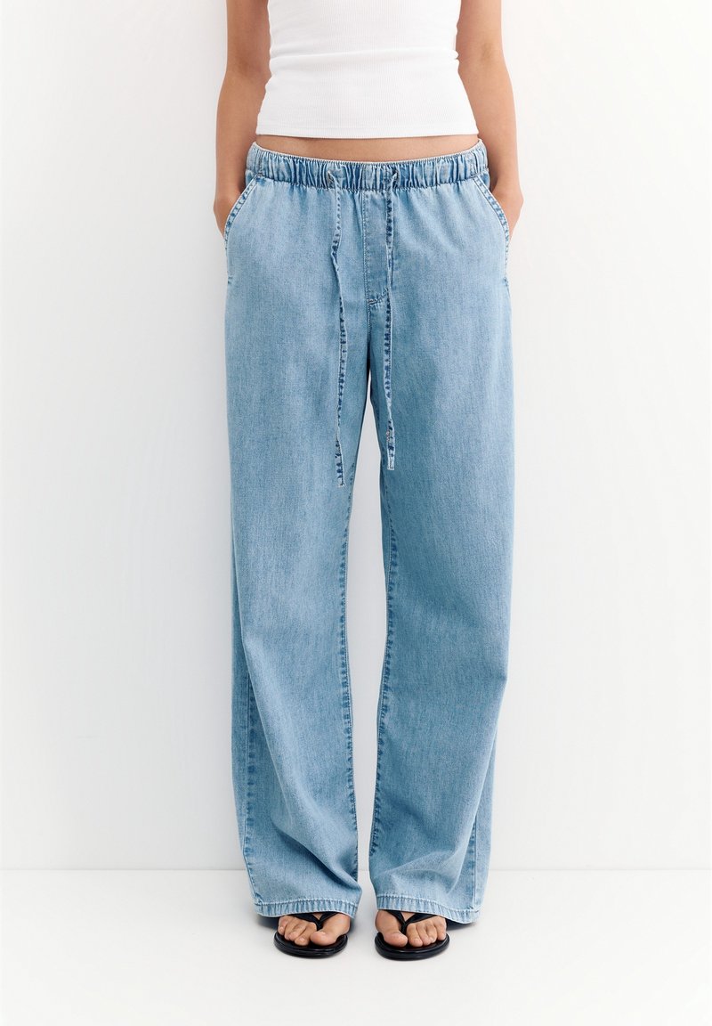 Personne portant un pantalon en jean bleu clair ample avec une taille élastique et un cordon, un débardeur blanc court, et des sandales noires flip-flop.