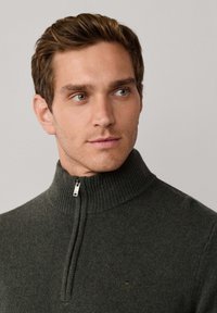 Donkergroene gebreide pull met rits en een geribde kraag, voorzien van een klein logo op de borst en een zachte textuur.