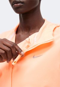 Άτομο με μπουφάν Nike σε χρώμα ροδάκινο τραβά ένα σφυρίχτρα που είναι προσαρτημένη στο φερμουάρ του μπουφάν κοντά στην κλείδα.