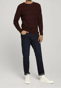 Gestrickter, kastanienfarbener Pullover mit langen Ärmeln, kombiniert mit dunkelblauen Jeans und weißen High-Top-Sneakern. Lässiges Outfit.