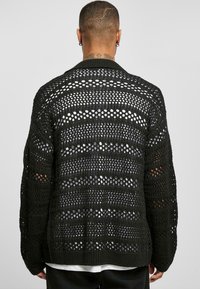 Juodas megztas kardiganas su laisvu, atviru audiniu, horizontaliomis juostelėmis ir įrėžtu apykakle. Ilgos rankovės, atsipalaidavęs siluetas.