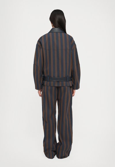 Costum rayat în albastru și maro, cu o croială relaxată, mâneci lungi și detaliu cu curea din piele. Textură subtilă pe material, cu pantaloni evazați.