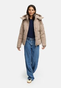 Beige Steppjacke mit Kapuze, ausgestattet mit einem Reißverschluss und Druckknopfverschluss, getragen über einem dunkelblauen Pullover und lockeren blauen Jeans. Legere Sneakers runden das Outfit ab.