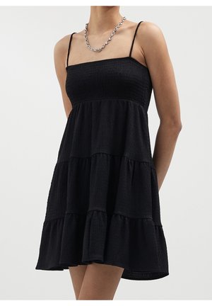 Kvinde iført sort, ærmeløs mini-kjole med lag og tynde stropper samt sølvkædehalskæde, stående foran en ensfarvet baggrund.