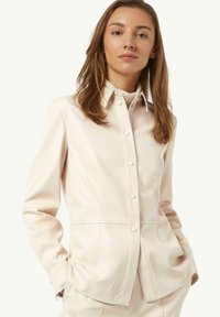 Cremfarbene Kunstlederhemdjacke mit durchgehender Knopfleiste, spitzem Kragen und Seitentaschen. Merkmale eine figurbetonte Silhouette und glatte Textur.