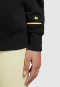 Czarna bluza z ribbowymi mankietami, z żółtym logo liścia oraz poziomą żółtą paskiem na rękawie. Miękka tekstura, casualowy design.