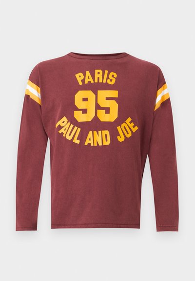 Chemise à manches longues bordeaux avec texte jaune "PARIS 95 PAUL AND JOE" et rayures blanches et jaunes sur le haut des manches.