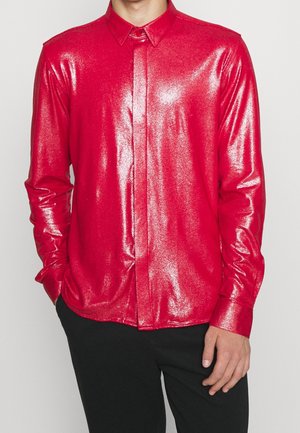 Mężczyzna ubrany w błyszczącą, metaliczną czerwoną koszulę z długim rękawem i kołnierzykiem, połączoną z czarnymi spodniami, stojący na jednolitym tle.
