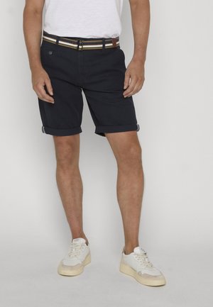 Mężczyzna w granatowych podwiniętych szortach, pasku w paski, białej koszulce i białych trampkach stoi na jednolitym tle.