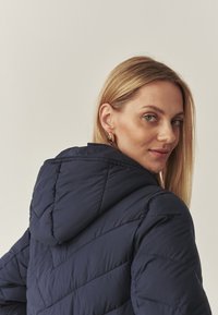 Tengeréskék puffos kabát kapucnival, steppelt mintázattal és puha tapintással. A modellen hosszú szőke haj van, és kerek fülbevalót visel.