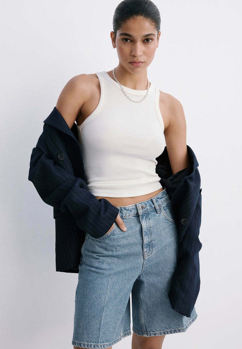 Femme portant un haut blanc sans manches, un short en jean bleu clair, un collier en chaîne argenté, avec une veste sombre posée sur ses épaules.