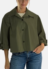 Olivgrön kort jacka i ett slätt tyg med stor krage och tre knappar. Har uppvikta ärmar med remdetaljer. Matchas med blå jeans.