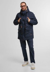Navy blue hooded parka with visible button closures, two front pockets, and contrasting brown accents. Worn over matching pants and sneakers.