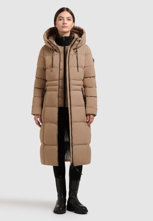 Woman wearing a long tan padded coat with hood, black pants, and black boots, standing against a plain light gray background.