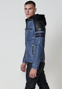Uomo che indossa una giacca blu con toppe trapuntate nere sulle spalle e sulle maniche, un cappuccio nero e pantaloni neri, in piedi di fronte a uno sfondo semplice.