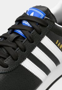 Fekete Adidas sportcipő fehér csíkokkal, fekete fűzőkkel, kék logóval a nyelven, és arany "SAM" felirattal a texturált bőr felsőrészen.