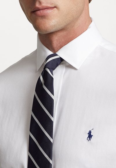White dress shirt with a herringbone pattern and a navy tie featuring white stripes. A small blue polo logo is visible on the shirt.