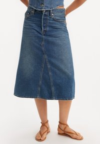 Denim midi skjørt i mellomblå, med en front søm, rett snitt og rå kant. Kombinert med lysebrune sandaler med stropper.