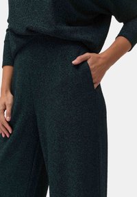 Femme portant un pantalon texturé foncé et un haut assorti, une main dans la poche et l'autre reposant le long du corps.