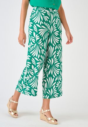 Femme portant un pantalon vert à jambes larges avec des motifs de feuilles blanches et des sandales compensées dorées, debout devant un fond uni.