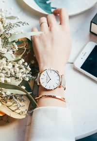 Orologio argento con quadrante bianco e dettagli in oro rosa su un polso, abbinato a un bracciale d'oro, circondato da fiori e uno smartphone.