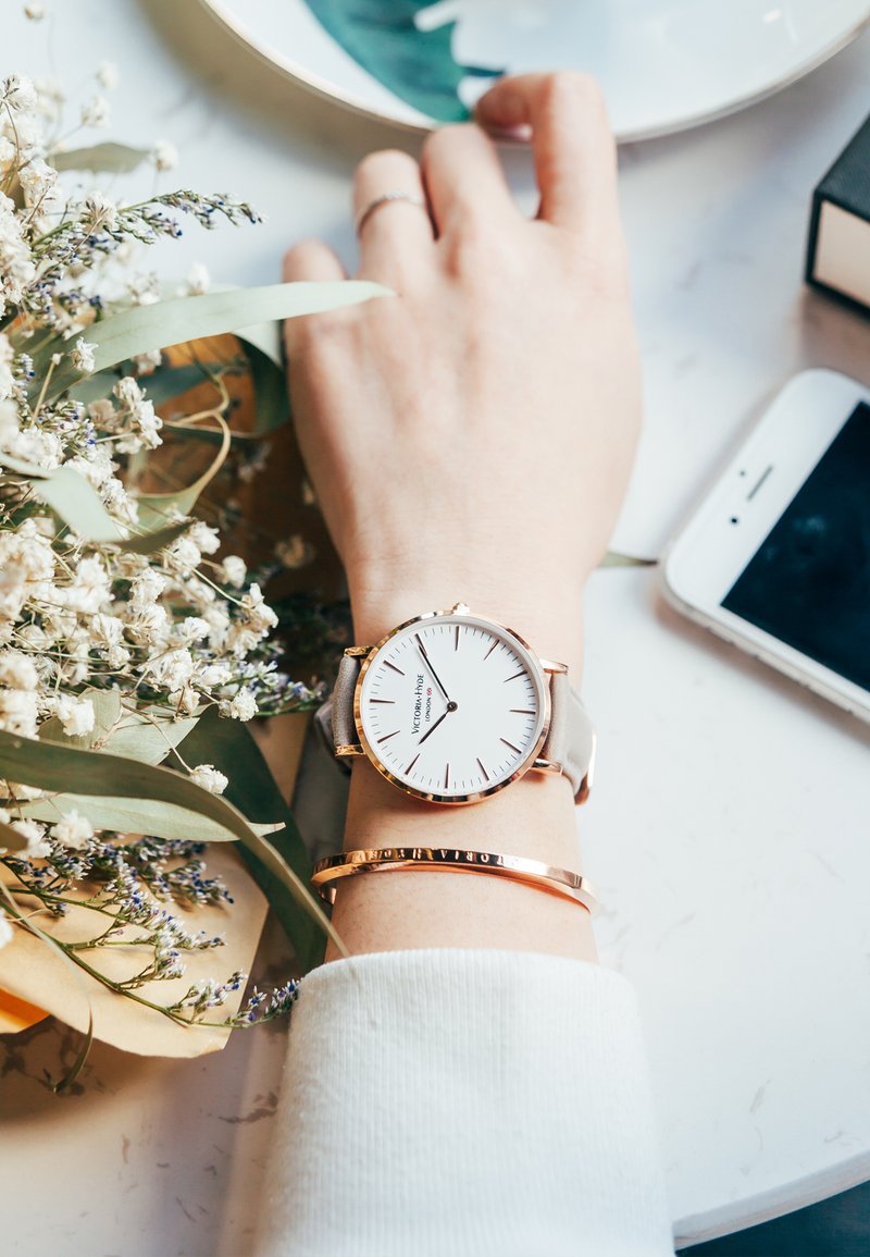 Orologio argento con quadrante bianco e dettagli in oro rosa su un polso, abbinato a un bracciale d'oro, circondato da fiori e uno smartphone.
