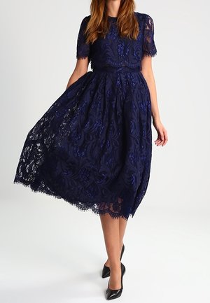 Frau trägt ein marineblaues Spitzen-Midikleid mit kurzen Ärmeln, hält den Rock und steht in schwarzen spitzen Schuhen.