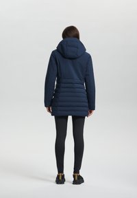 Marineblauwe pufferjack met capuchon, met een verticale gestikte textuur, een getailleerde taille en een iets verlengd zoom. Gecombineerd met zwarte leggings.