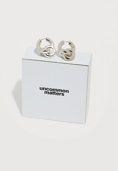 Ασημένια σκουλαρίκια σε σχήμα στριφογυριστού κρίκου, τοποθετημένα σε λευκό κουτί με την επιγραφή "uncommon matters" σε απλό φωτεινό φόντο.