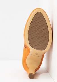 Chaussure à talon en daim orange reposant sur le côté, montrant la semelle beige avec une surface adhérente marron texturée et la taille 37 marquée.