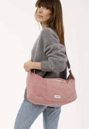 Rosa Stoff-Schultertasche mit einem Reißverschlussfach, weicher Textur und kontrastierendem braunem Trageriemen, die von einer Person in einem grauen Pullover gehalten wird.