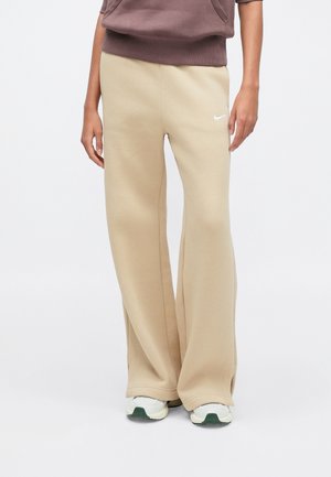 Personne portant un pantalon de survêtement beige Nike à jambes larges et des baskets blanches, visible de la moitié du torse aux pieds sur un fond uni.