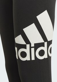 Leggings negros hechos de una tela suave y elástica que presentan un gran logo blanco de Adidas y tres rayas diagonales en el lateral.