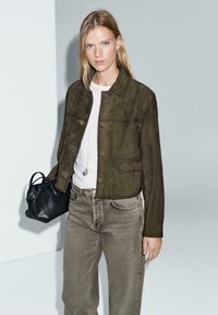 Giacca in suede verde oliva con colletto e bottoni frontali, abbinata a una camicia chiara e jeans beige a vita alta; tiene in mano una piccola borsa nera.