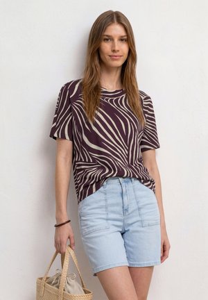 Femme portant une chemise à motifs violets et beiges avec un short bleu clair, tenant un sac en paille tressée devant un mur blanc uni.