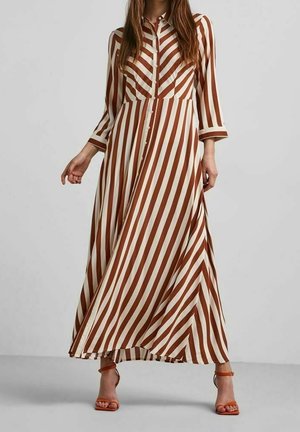 Mujer que lleva un vestido largo abotonado con rayas diagonales y verticales en marrón y blanco, combinado con tacones marrones de punta abierta.