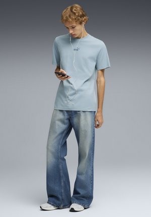 Giovane che indossa una t-shirt azzurro chiaro Puma e jeans sbiaditi a gamba larga, che guarda uno smartphone con gli auricolari bianchi collegati.