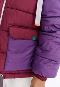 Kurtka puchowa z panelami w kolorze bordowym i purpurowym, z zielonym akcentem na naszywce kieszeni i białym suwakiem z pomarańczowym podkreśleniem.