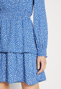 Robe bleue à pois blancs, avec des manches bouffantes, un corsage smocké et un design de jupe superposée. Texture de tissu léger.