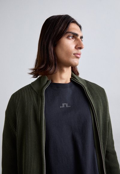 Jauns cilvēks ar plecu garuma tumšiem matiem, ģērbies melnā krekliņā un atvērtā tumši zaļā ribotā zip-up džemorā, skatās pa labi.