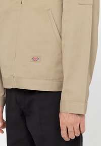 Beige Dickies-jacka på en person som bär svarta byxor, visar framlappen, manschettdetaljer och en liten handtatuering.