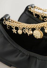 Chaussures en cuir noir avec une chaîne dorée ornée de breloques en forme d'étoile, de cercle, de nœud et de disque texturé sur le devant.