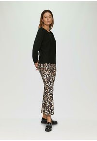 Eine Frau steht da und trägt einen schwarzen Pullover, Leopard-Print-Hosen und schwarze Loafers vor einem schlichten hellen Hintergrund.