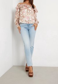 Mujer con jeans claros, blusa floral sin hombros, tacones de plataforma marrones, de pie contra una pared blanca en la esquina con las manos en las caderas.