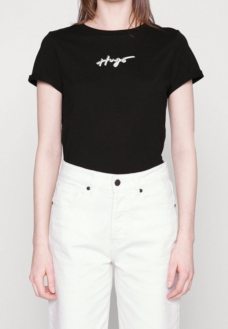 Femme portant un T-shirt noir avec "hugs" écrit dessus en lettres blanches et un jean blanc taille haute, debout devant un fond uni.