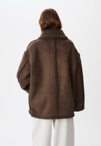 Giacca in shearling sintetico marrone con colletto alto, vestibilità oversize e dettaglio della fibbia laterale. Superficie testurizzata con finitura morbida, senza motivi visibili.