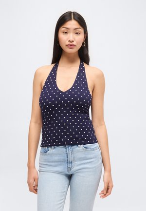 Kobieta ubrana w granatowy top bez rękawów z białymi grochami i jasnoniebieskie dżinsy, stojąca na tle jednolitego jasnego tła.