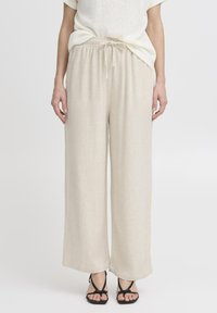 Pantalons larges beige en tissu léger avec une taille �à cordon de serrage et une surface lisse légèrement texturée. Assortis avec des sandales noires.