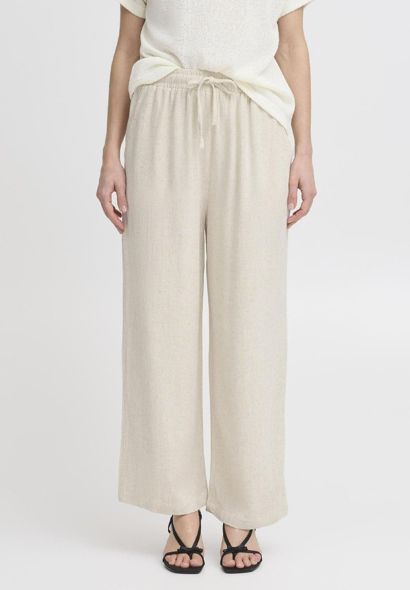 Pantalons larges beige en tissu léger avec une taille à cordon de serrage et une surface lisse légèrement texturée. Assortis avec des sandales noires.