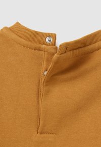 Top color mostaza con cuello acanalado, que presenta dos botones a presión en la parte posterior. La tela es suave y de textura lisa.